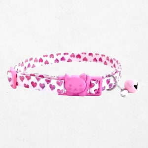 May include: A pet collar featuring a white fabric strap with a repeating pattern of pink hearts. The collar includes a pink plastic buckle shaped like a cat head, a metal D-ring, and a small pink bell attached to a silver-toned metal ring.