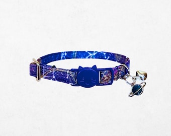 Puede incluir: Un collar azul para gatos con estampado de galaxia. El collar presenta una hebilla azul con forma de gato y una campana plateada con un dije de planeta. El collar mide aproximadamente 1,3 cm de ancho.