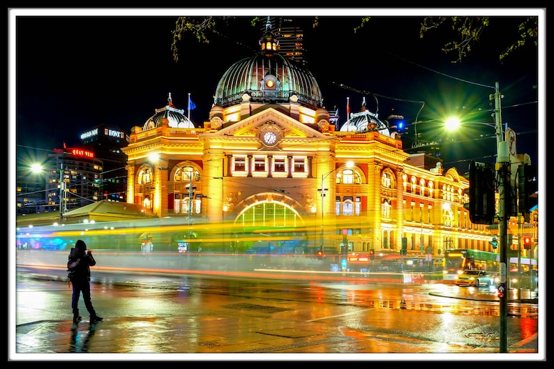 Pu&ograve; includere: Una vista notturna di un grande edificio ornato con una torre dell'orologio. L'edificio &egrave; illuminato da luci gialle calde, e in primo piano ci sono auto e lampioni. L'edificio si riflette sul selciato bagnato.