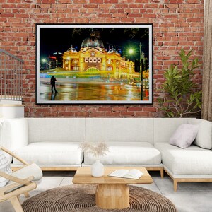 Pu&ograve; includere: Una fotografia incorniciata di un grande edificio illuminato con una torre dell'orologio di notte. L'edificio si riflette in una strada bagnata, e c'&egrave; una persona che cammina in primo piano.