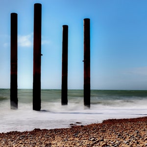 May include: Four tall, weathered, dark brown wooden posts stand in the surf on a sandy beach. The ocean waves are blurred, creating a sense of motion. The sky is a clear blue.