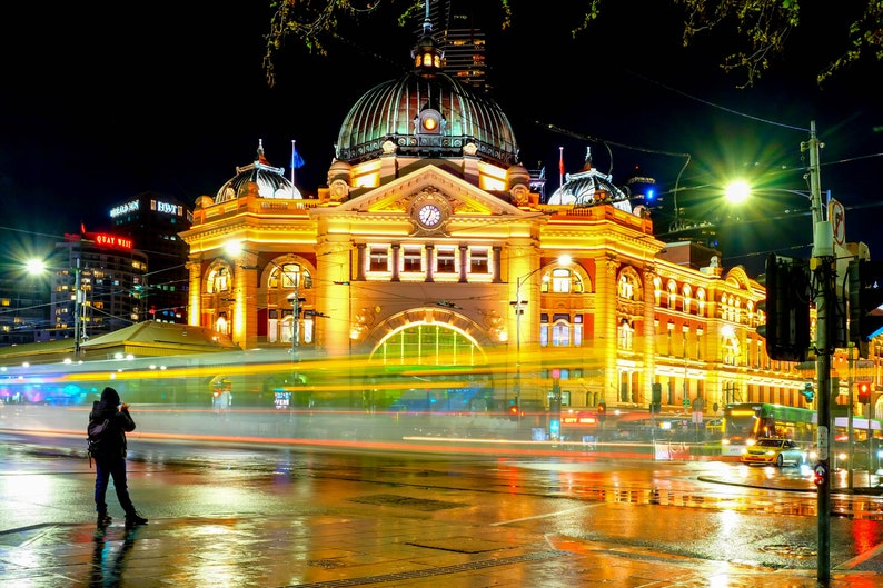 Pu&ograve; includere: Una vista notturna di un grande edificio ornato con una torre dell'orologio. L'edificio &egrave; illuminato da luci gialle calde, e in primo piano ci sono auto e lampioni. L'edificio si riflette sul selciato bagnato.