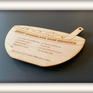 May include: A wooden bowl-shaped cookie cutter with a recipe for chocolate chip cookies engraved on it. The recipe includes ingredients like butter, brown sugar, milk, flour, chocolate chips, baking soda, salt, and vanilla. The cutter also has a small wooden spoon attached to the side.