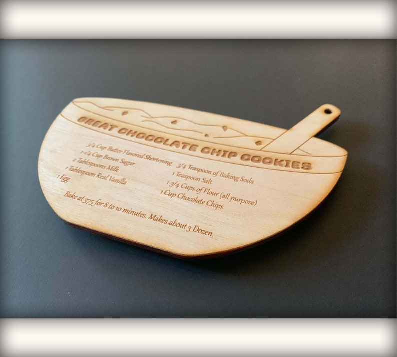 May include: A wooden bowl-shaped cookie cutter with a recipe for chocolate chip cookies engraved on it. The recipe includes ingredients like butter, brown sugar, milk, flour, chocolate chips, baking soda, salt, and vanilla. The cutter also has a small wooden spoon attached to the side.