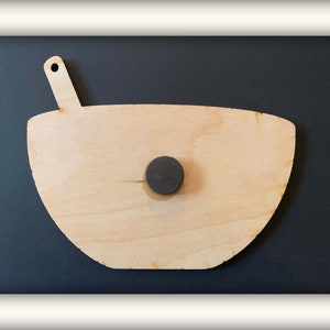 May include: A wooden bowl with a spoon attached to the side. The bowl is light brown and has a natural wood grain. A black magnet is attached to the back of the bowl.