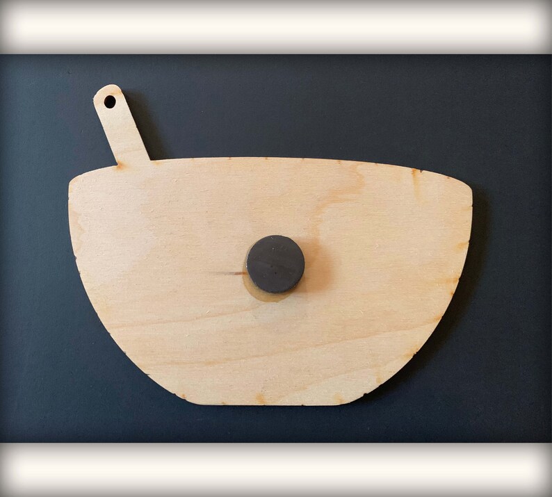 May include: A wooden bowl with a spoon attached to the side. The bowl is light brown and has a natural wood grain. A black magnet is attached to the back of the bowl.
