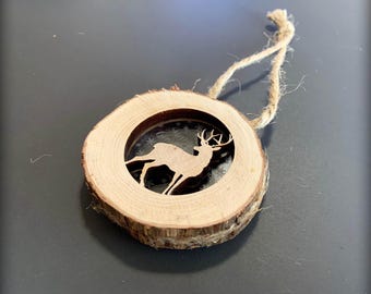 May include: A wooden ornament shaped like a tree trunk with a cutout of a deer inside. The ornament has a rustic brown color and is attached to a twine string.