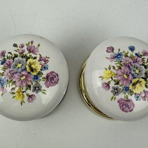 May include: Two white, round compacts with floral designs. Each compact features a bouquet of purple, pink, yellow, and blue flowers. One compact has a gold-coloured rim.