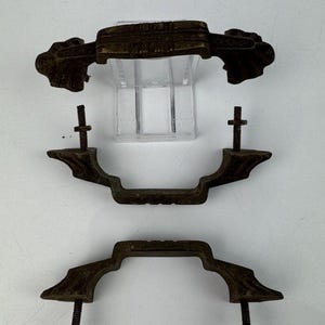 Könnte beinhalten: Drei antike Schubladengriffe in Bronzetönen mit aufwendigen Designs. Jeder Griff hat einen gebogenen Griff und zwei Gewindestifte zur Befestigung. Die Griffe werden vor einem weißen Hintergrund präsentiert, der ihr gealtertes Aussehen und ihre komplizierten Details hervorhebt.