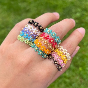 May include: A set of colourful daisy flower beaded rings. The rings are made with small, colourful beads that are arranged in a daisy flower pattern. The rings are stacked on top of each other, with the colours ranging from black to white to pink to yellow to green to blue.