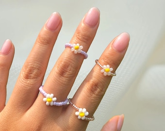 May include: Four dainty rings with a daisy design. Each ring is made of small white beads with a yellow center. The rings are worn on different fingers of a hand.