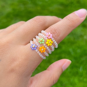 May include: Five colourful beaded rings with flower charms. The rings are stacked on a finger and are made with clear beads and small flower charms in purple, green, yellow, orange, and pink.
