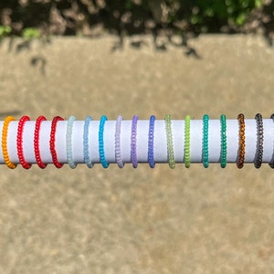 May include: A row of 15 colorful beaded rings on a white surface. The rings are arranged in a rainbow pattern, starting with orange and ending with black.