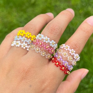 May include: A set of five beaded rings featuring daisy-shaped beads in various colours, including yellow, white, pink, purple, and red. The rings are stacked on a finger, showcasing the different colours and designs.