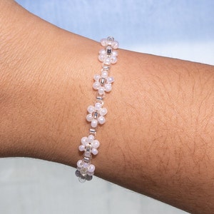 May include: A white beaded bracelet with small flower-shaped charms. The bracelet is made with white beads and silver accents.
