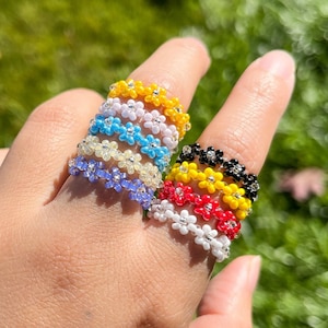 May include: A set of seven colourful beaded rings, each featuring a flower design. The rings are stacked on a finger, with the colours ranging from yellow to blue to red.