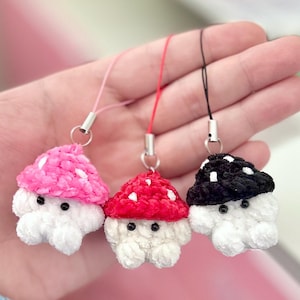 May include: Three crochet mushroom charms in pink, red, and black with white spots. Each mushroom has a white base and small black eyes. The charms are attached to silver-colored metal loops with pink, red, and black cords.
