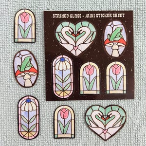 Pode incluir: Uma folha de autocolantes com o título STAINED GLASS - MINI STICKER SHEET apresentando vários desenhos, incluindo dois cisnes formando um coração, um sapo num cogumelo e várias janelas em arco com túlipas e margaridas.