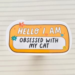 Puede incluir: Una pegatina amarilla con esquinas redondeadas y borde blanco. La pegatina tiene un fondo blanco con un rectángulo amarillo en el centro. El texto "HELLO I AM OBSESSED WITH MY CAT" está escrito en negro sobre el rectángulo amarillo. La pegatina tiene una carita sonriente blanca en la esquina inferior derecha.