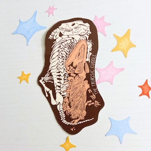 Puede incluir: Una pegatina marrón con un esqueleto de dinosaurio y una criatura peluda, con el texto "THE TRIASSIC CUDDLE". Alrededor de la pegatina hay recortes en forma de estrella de colores azul, rosa, naranja y amarillo. Sobre una superficie blanca.