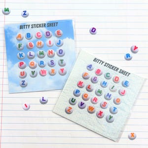 May include: Two "Bitty Sticker Sheet" packs with colorful alphabet stickers. Each sticker is a small, round shape with a letter. The background of one sheet has a sky and cloud design. Individual stickers are scattered around.