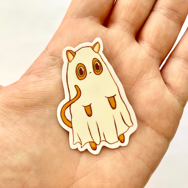 May include: A die-cut sticker featuring a cartoon cat dressed as a ghost. The cat is white with tan ears, tail, and paws. It has large, round eyes with brown irises. The ghost costume is a cream color.