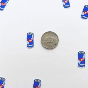 Puede incluir: Una colecci&oacute;n de peque&ntilde;as pegatinas de latas de Red Bull azules y blancas con un logotipo rojo, dispersas alrededor de una moneda de n&iacute;quel sobre un fondo blanco. Las pegatinas est&aacute;n en varias orientaciones.