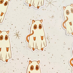 May include: A collection of stickers featuring a cartoon cat dressed as a ghost. The cat is white with orange ears, eyes, and tail. The ghost costume has a draped appearance. The background is white with gold flecks and star designs.