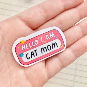 May include: A pink and white sticker with the text "Hello I am Cat Mom" in black lettering. The sticker has a white border and is decorated with small yellow and blue flowers.