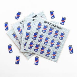 Puede incluir: Hojas de pegatinas y pegatinas individuales con un dise&ntilde;o de lata de Red Bull. Las hojas de pegatinas son de color azul claro con m&uacute;ltiples pegatinas de latas de Red Bull. Las pegatinas individuales est&aacute;n dispersas. El texto "Bitty Sticker Sheet" es visible.