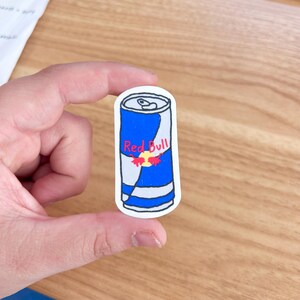 May include: A white sticker with a hand-drawn illustration of a blue and white Red Bull can. The can has the Red Bull logo and two red bulls on the front.