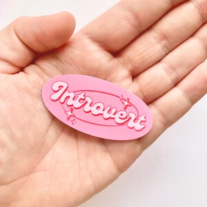 May include: Pink oval sticker with the word "Introvert" in white letters with a pink outline. The sticker has a white star design around the word.