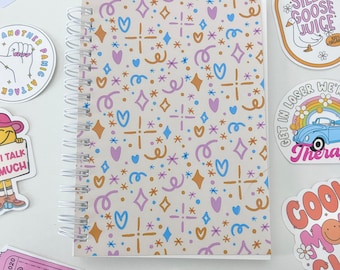 May include: A spiral bound notebook with a beige cover featuring a repeating pattern of hearts, stars, and swirls in shades of pink, blue, and gold. The notebook is surrounded by various colorful stickers with humorous and quirky phrases.