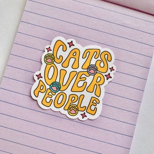 May include: A yellow sticker with the text "Cats Over People" in a retro font. The sticker is decorated with paw prints in pink, blue, and yellow.