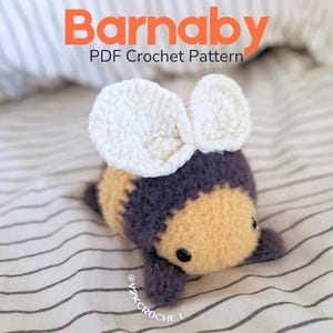 Peut inclure: Un jouet d'abeille au crochet avec des ailes blanches, un corps jaune et une tête et des pattes violettes. Le texte "Barnaby PDF Crochet Pattern" est affiché en orange. L'abeille est posée sur une surface rayée blanche et grise.