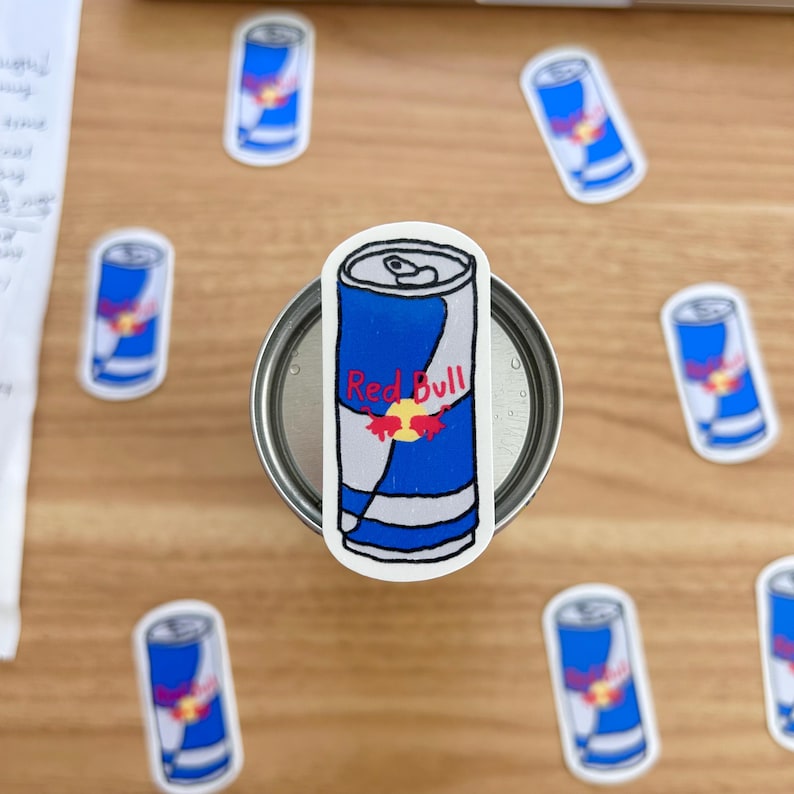 May include: A set of eight red bull stickers, each featuring a blue and white can with the red bull logo and the words "Red Bull" in red. The stickers are on a wooden surface.