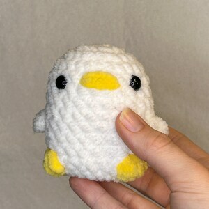 May include: A white crocheted duck with yellow beak and feet. The duck has black eyes.