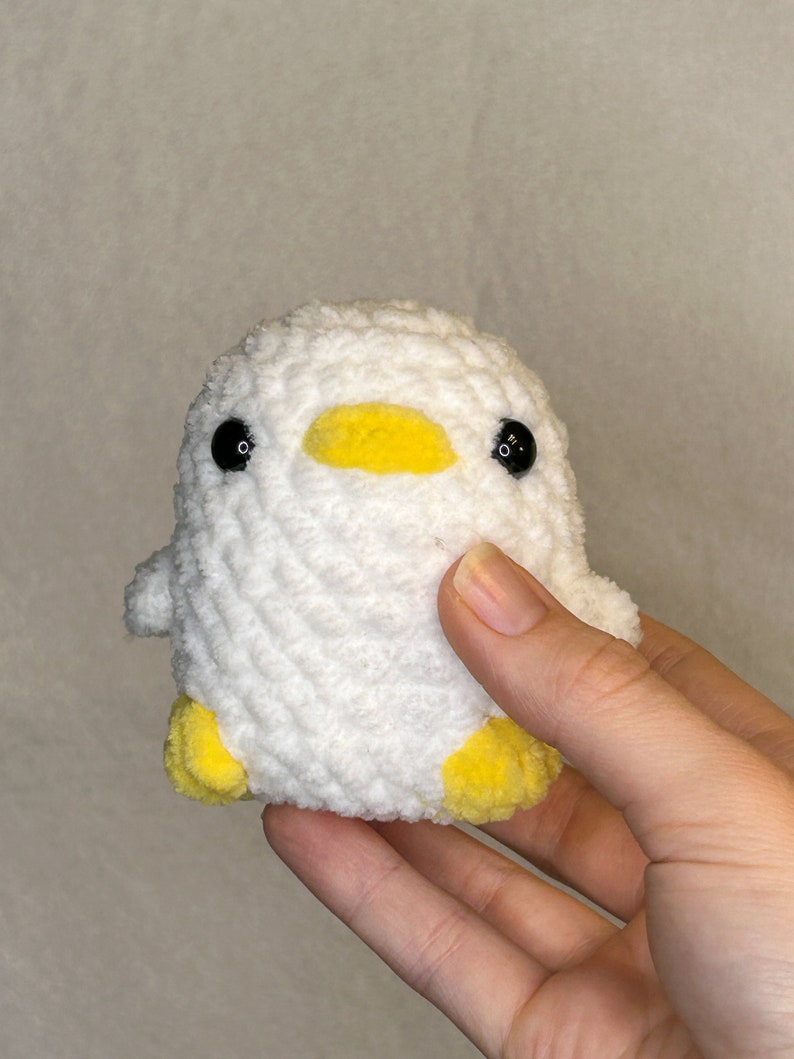 May include: A white crocheted duck with yellow beak and feet. The duck has black eyes.