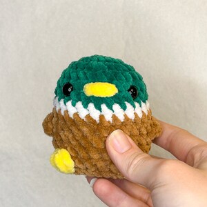 Peut inclure: Un jouet de canard en crochet avec une t&ecirc;te verte, un bec jaune et un corps marron. La poitrine du canard est blanche et les pieds sont jaunes. Le jouet est fait de fil doux et textur&eacute; et a des yeux noirs en forme de bouton.