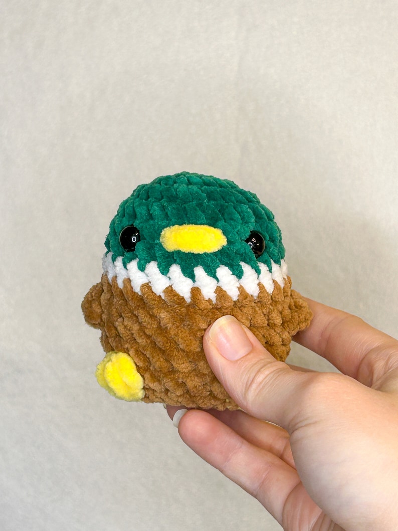 Peut inclure: Un jouet de canard en crochet avec une t&ecirc;te verte, un bec jaune et un corps marron. La poitrine du canard est blanche et les pieds sont jaunes. Le jouet est fait de fil doux et textur&eacute; et a des yeux noirs en forme de bouton.