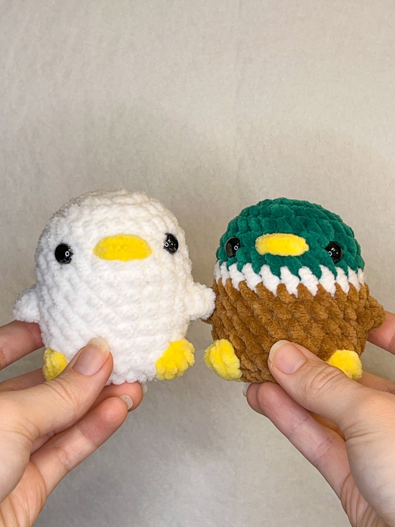 Peut inclure: Deux canetons crochet&eacute;s &agrave; la main. L'un est blanc avec un bec et des pieds jaunes, l'autre est marron, vert et blanc avec un bec et des pieds jaunes. Les deux ont des yeux de s&eacute;curit&eacute; noirs. Les canetons sont petits et en peluche.