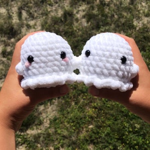 May include: Two white, crocheted ghost-shaped plush toys with black eyes and pink cheeks are held up against a blurred green and brown background. The plush toys are joined at the base, appearing to be kissing.