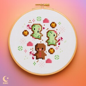 May include: A cross-stitch embroidery hoop featuring a festive design. The design includes three gingerbread-style creatures, two green and one brown, surrounded by candy canes, cookies, and pink and green decorative elements. The hoop is set against a gradient background.