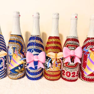 May include: Six glass bottles covered in colorful rhinestones, featuring various designs including sports logos, animal graphics, and the year 2025. Each bottle has a white foil-wrapped neck and is decorated with sparkling gems in blue, gold, red, and white.