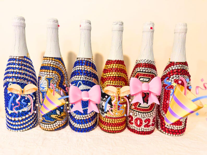May include: Six glass bottles covered in colorful rhinestones, featuring various designs including sports logos, animal graphics, and the year 2025. Each bottle has a white foil-wrapped neck and is decorated with sparkling gems in blue, gold, red, and white.