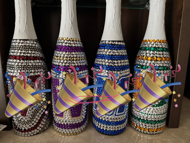 May include: Four glass bottles with white foil-wrapped necks, each fully covered in rows of sparkling rhinestones in red, purple, blue, and green patterns. Each bottle is partially obscured by a gold and purple party popper graphic.