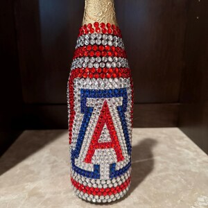 May include: A glass bottle covered in red, white, and blue rhinestones. The neck is wrapped in gold foil. The body features a large letter A in red, white, and blue rhinestones, designed to resemble the University of Arizona logo emblem.