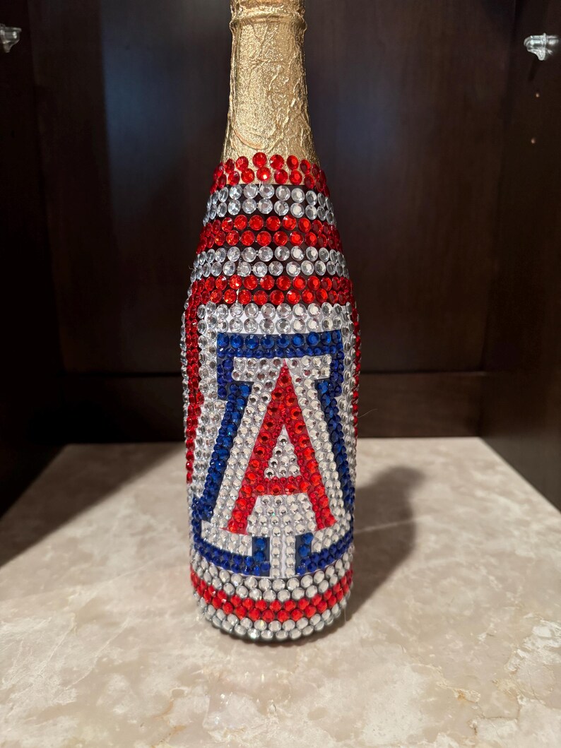 May include: A glass bottle covered in red, white, and blue rhinestones. The neck is wrapped in gold foil. The body features a large letter A in red, white, and blue rhinestones, designed to resemble the University of Arizona logo emblem.