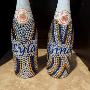 May include: Two Martinelli's sparkling juice bottles decorated with clear, blue, and gold rhinestones in a wavy pattern. The left bottle features the name Lyla in blue script, and the right bottle features the name Gina in blue script.