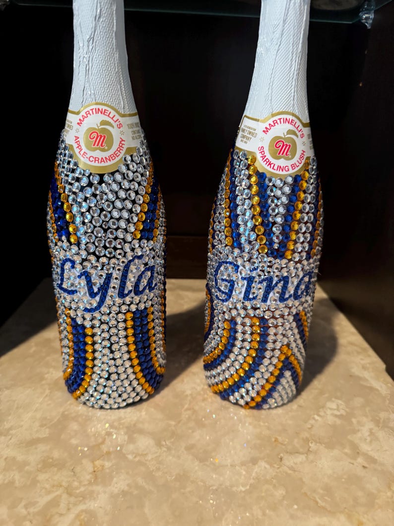 May include: Two Martinelli's sparkling juice bottles decorated with clear, blue, and gold rhinestones in a wavy pattern. The left bottle features the name Lyla in blue script, and the right bottle features the name Gina in blue script.